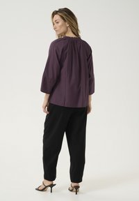 Lila, leichte Bluse mit gerafftem Ausschnitt, Dreiviertelärmeln und lockerer Passform, kombiniert mit schwarzen, schmal zulaufenden Hosen und High Heels.
