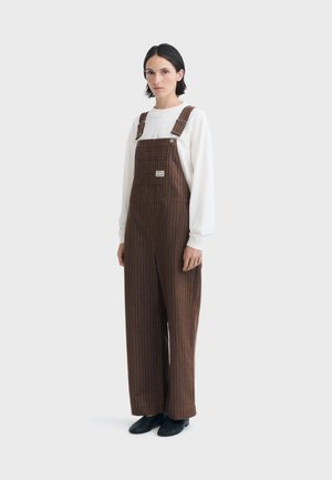 Femme aux cheveux courts et foncés portant une chemise blanche à manches longues et une salopette marron à fines rayures, debout devant un fond uni.