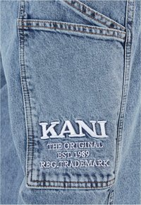 Šviesiai mėlyni džinsai su užpakaline kišenėle, kurioje balta siuvinėta frazė: "KANI ORIGINALAS EST 1989 REG. PREKĖS ŽENKLAS."