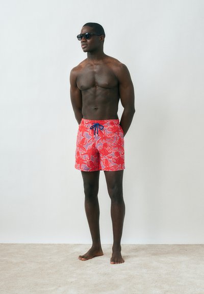 Μυώδης άνδρας με γυαλιά ηλίου και κόκκινα μαγιό με σχέδιο, στέκεται ξυπόλητος μπροστά σε απλό φωτεινό φόντο, με τα χέρια πίσω από την πλάτη.