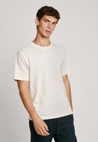 T-shirt leggero crema a maniche corte in cotone, con colletto rotondo e un logo discreto sul petto. Tessuto morbido e vestibilità rilassata.