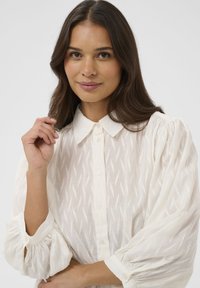 Blouse blanche boutonnée avec un motif texturé, dotée d'un col et de manches bouffantes. Tissu doux avec une apparence légère et transparente.