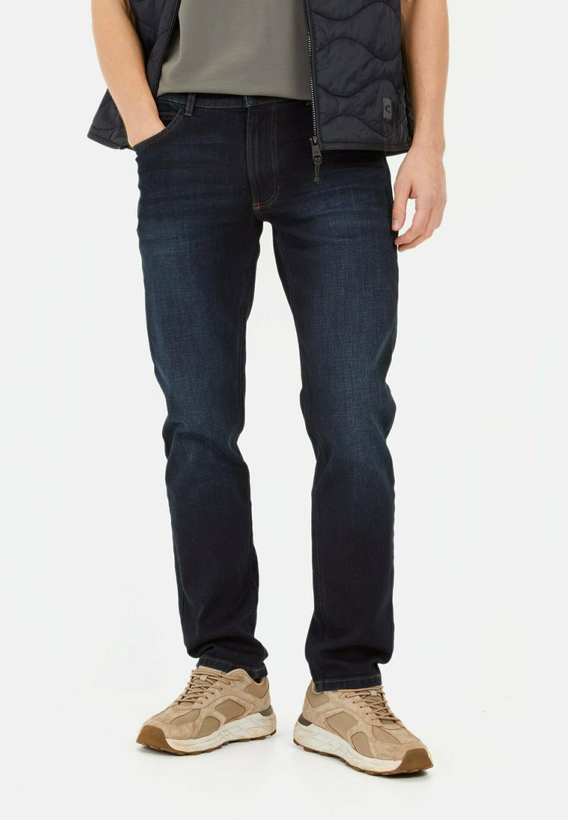 Donkerblauwe jeans met een rechte snit, gemaakt van denim. Gepaard met lichtbruine sneakers en een grijze shirt, gedragen onder een zwarte puffervest.