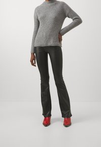 Persona vestita con un maglione grigio a coste, pantaloni di pelle neri e stivaletti con tacco tempestati di borchie rossi, in piedi con una mano sulla vita.