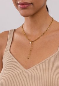 Goldene Kettenhalskette mit länglichen Gliedern und einem Tropfenanhänger mit einer kleinen Perle, getragen mit einem beigen gerippten Oberteil.