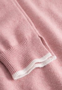 Jasnoróżowy sweter z detalem mankietu ściągacza z białym wykończeniem. Tekstura wydaje się miękka i lekko puszysta.