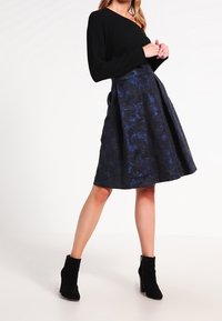 Svart långärmad topp i kombination med en knälång, texturerad kjol med ett blommönster i blått. Modellen bär svarta ankelboots.