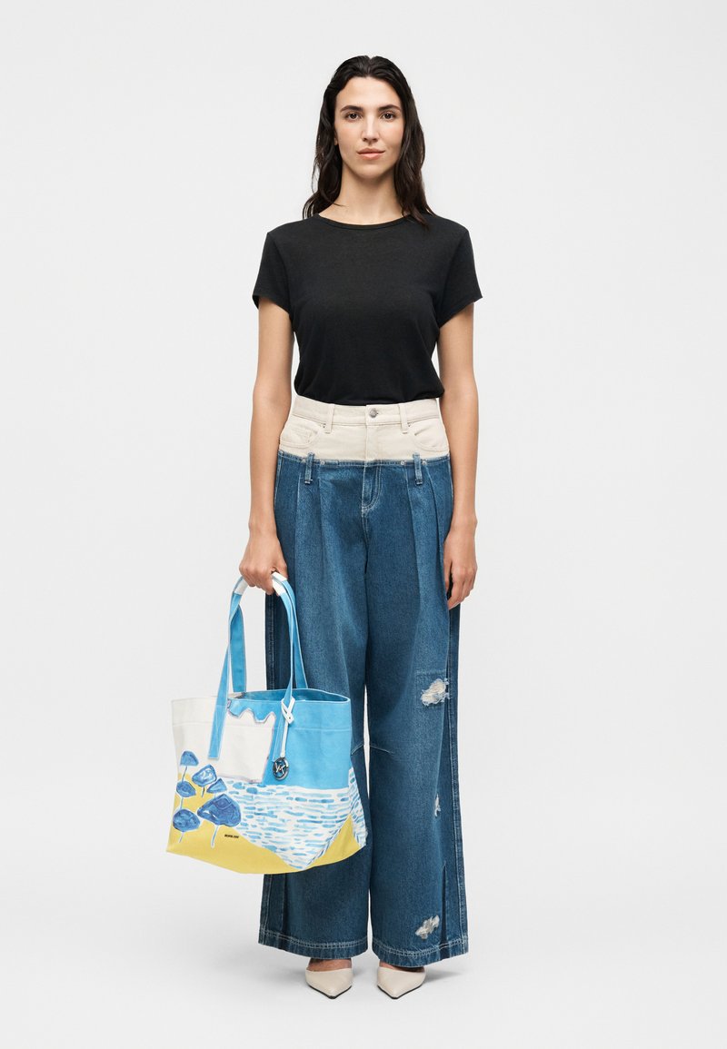 Femme debout portant un t-shirt noir et un jean large en denim, tenant un grand sac cabas avec un design abstrait bleu, jaune et blanc.