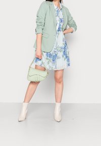 Mintgrön kavaj över en ljusblå blommig klänning. Modellen håller en mintgrön handväska. Beiga anklarstövlar fulländar outfiten.