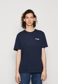 Temno modra bombažna majica s kratkimi rokavi in okroglim izrezom, s belim logotipom "Wrangler" na levi strani prsnega koša.