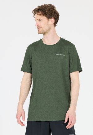 Mann mit heller Haut und lockigem braunem Haar trägt ein grünes Kurzarmshirt und schwarze Shorts und blickt nach links vor weißem Hintergrund.