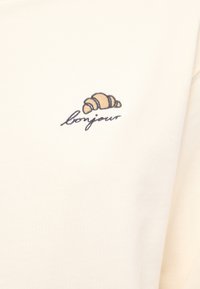 Felpa color crema con un croissant ricamato e la parola "bonjour" in corsivo nero sotto il design. Tessuto morbido e texturizzato.