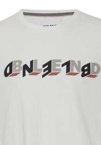 Hvid T-shirt med fed, stiliseret sort og brun 3D-tekstdesign tværs over brystet, der indeholder en blanding af solide og prikkede mønstre.