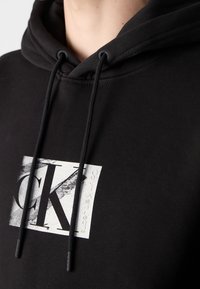 Sudadera negra con cordones y un logotipo rectangular blanco de Calvin Klein Jeans que presenta grandes letras "CK" en el pecho.