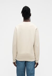 Béžová mikina s okrúhlym výstrihom a rebrovanými manžetami, vyrobená z mäkkého materiálu. Na sebe modré džínsové nohavice, vidieť zozadu.