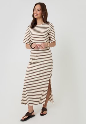 Frau trägt ein creme- und braun gestreiftes Midikleid mit kurzen Ärmeln und Seitenschlitz, lächelt und steht vor einem schlichten hellen Hintergrund.