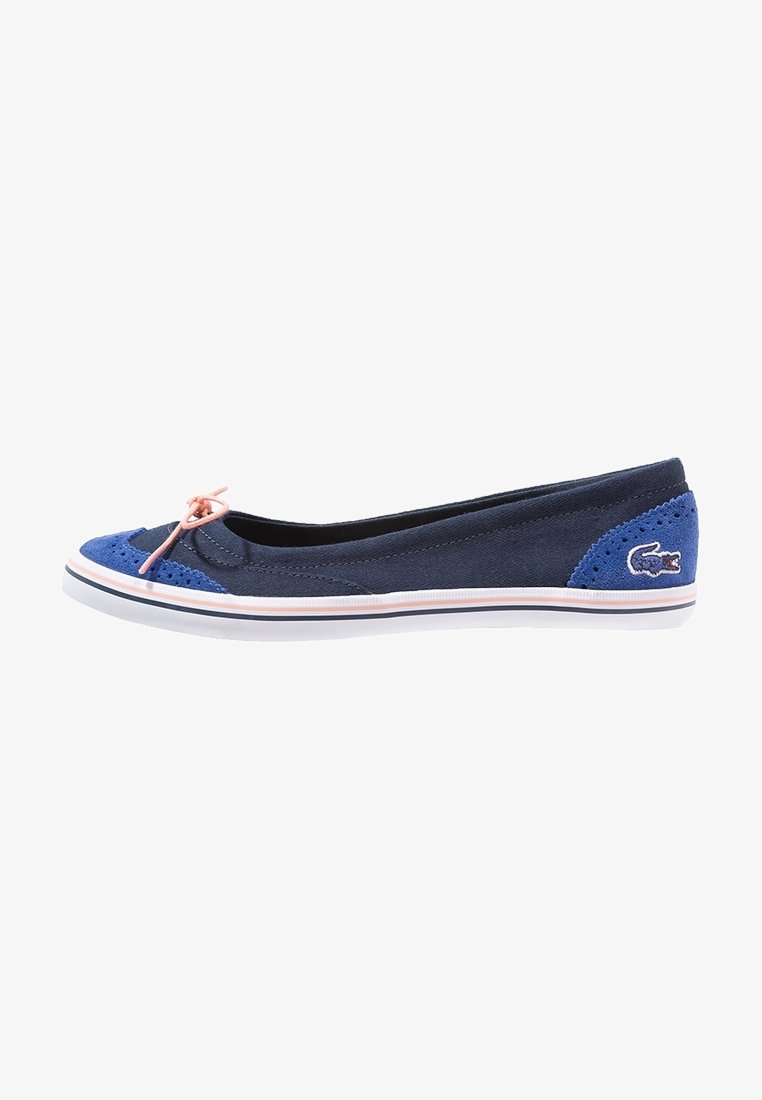 Ballerine en toile bleu marine avec des accents en daim bleu clair, nœud rose sur le bout, semelle en caoutchouc blanche et petit logo crocodile brodé près du talon.