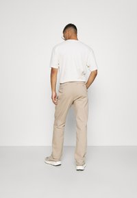 Beige cotton pants with a straight-leg design, featuring back pockets and a subtle logo detail. Paired with a white short-sleeve shirt.
