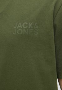 Camiseta de manga corta verde oscuro, hecha de una tela suave, con un logotipo en relieve en tono sobre tono que dice "JACK & JONES" en el frente.