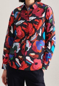 Chemise boutonnée à manches longues colorée, avec un fond sombre et de grands motifs floraux en rouge, bleu, vert et crème. Texture de tissu lisse.