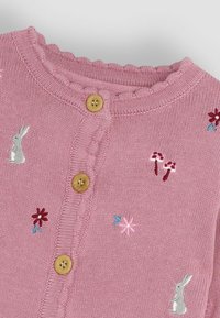 Rozā adīta kardigana ar viļņu apakšmalu, koka pogām un izšūtiem motīviem ar trušiem un ziediem sarkanā, zilā un sudraba krāsā.