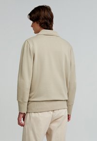 Beige sweatshirt with a collared neckline, ribbed cuffs, and a textured finish. Worn with light-colored pants, viewed from the back.