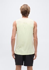 Homme aux cheveux blonds courts portant un débardeur jaune clair et un short noir, debout face à un fond blanc.