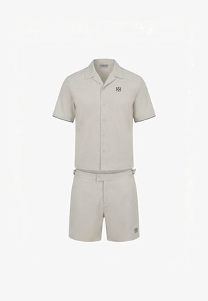 Homme portant une chemise beige clair à manches courtes boutonnée et un short assorti avec des sandales, debout devant un fond blanc uni.