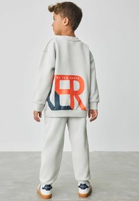 Helles graues Sweatshirt und Hose mit einem großen roten und blauen Grafikmotiv auf dem Rücken. Weicher Stoff und entspannte Passform. Sneaker mit kontrastierendem Logo.