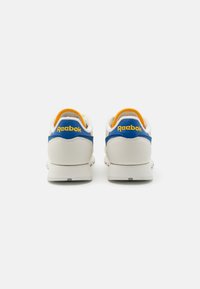 Białe trampki z niebieskimi i żółtymi akcentami, z teksturowanym obcasem, okrągłym kształtem i gładkim wykończeniem. Logo "Reebok" z tyłu.
