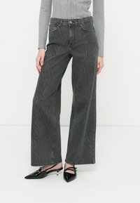 Jeans de mezclilla negras de tiro alto y pernera ancha con un sutil patrón de ondas y cierre de botón. Combinados con zapatos negros brillantes de punta afilada.