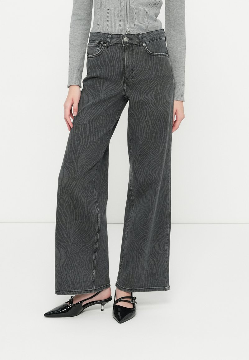 Jeans de mezclilla negras de tiro alto y pernera ancha con un sutil patrón de ondas y cierre de botón. Combinados con zapatos negros brillantes de punta afilada.