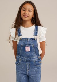 Los overoles de mezclilla presentan un lavado azul con patrones circulares y un parche rosa. Se combinan con una camiseta blanca con mangas acampanadas.