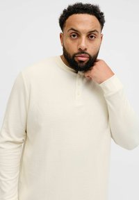 Kremfarget henley-skjorte med lange ermer og ribbestruktur, med rund hals og tre knapper nær kragen.