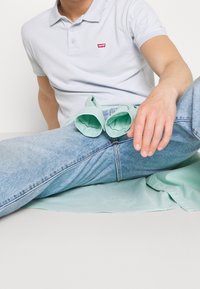 Světle modrá polokošile s malým červeným logem Levi's. Světle džínové kalhoty s volným střihem a mátově zelená košile přehozená přes klín.