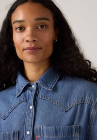 Denimskjorte med tryknapkrave, to brystlommer og kontrastsyninger. Lys blå med struktureret stof og synlige sømme.