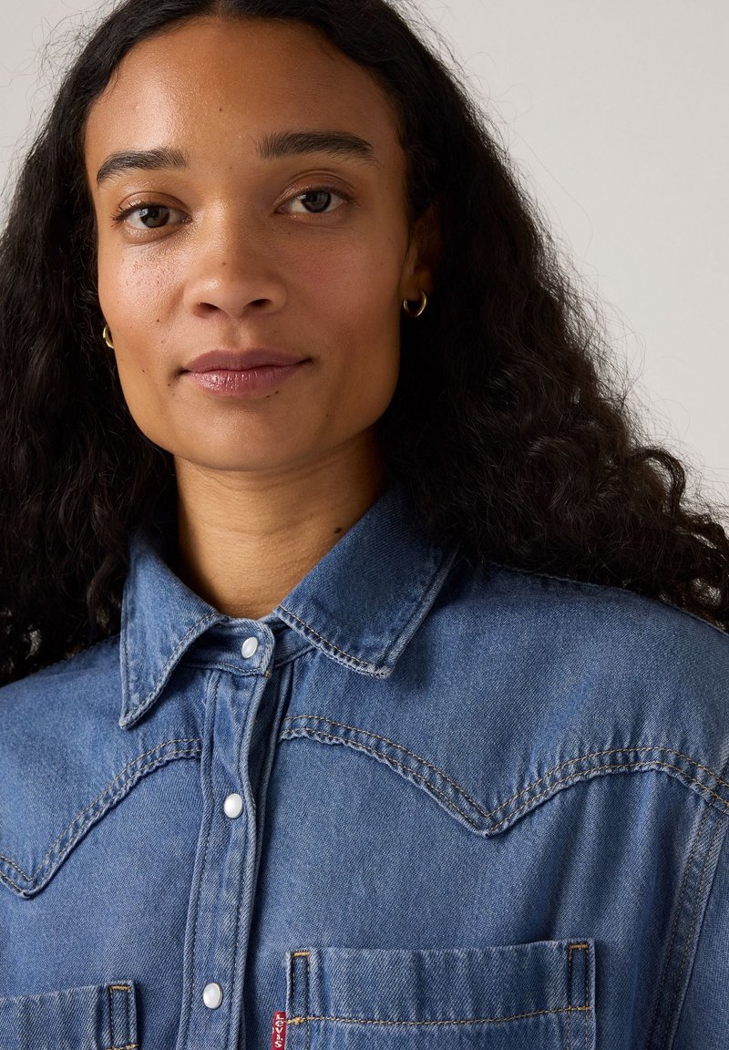 Denim overhemd met een drukknoopkraag, twee borstzakken en contrasterende stiksels. Lichtblauw met een gestructureerde stof en zichtbare naden.