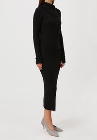 Robe en tricot noir côtelé avec un col montant, des manches longues et une fente sur le côté, associée à des escarpins gris clair à bout pointu.