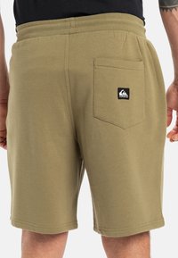 Olive green shorts made of soft cotton. Features an elastic waistband, rear pocket with a black logo patch, and hemmed cuffs.