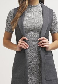 Harmaa hihaton neuletakki huivikaarteella, pehmeästä kankaasta, siinä on kaksi etutaskua, kerroksittain teksturoidun harmaan mekon päällä.