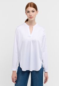 Camicia bianca a maniche lunghe con scollo a V e spacchi laterali, realizzata in tessuto morbido; indossata con pantaloni in denim blu, caratterizzati da una vestibilità comoda.