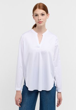Camicia bianca a maniche lunghe con scollo a V e spacchi laterali, realizzata in tessuto morbido; indossata con pantaloni in denim blu, caratterizzati da una vestibilità comoda.