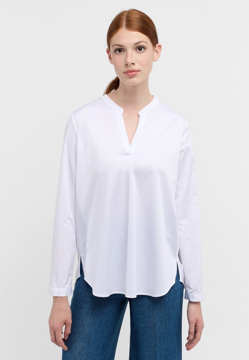 Witte lange mouwen shirt met een V-hals en zijsplitten, gemaakt van gladde stof; gedragen met blauwe denim broeken met een relaxte pasvorm.