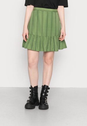 Saia verde com riscas, com uma bainha ondulada, feita de um tecido leve. Combinada com botas pretas, robustas, de atacadores, com várias tiras.