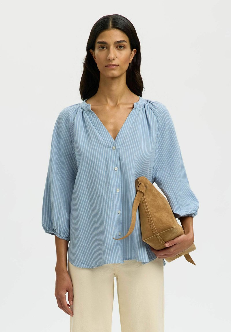 Femme aux cheveux longs et foncés portant un chemisier à rayures bleu clair et un pantalon crème, tenant un sac à dos en daim couleur tan sur un fond blanc.