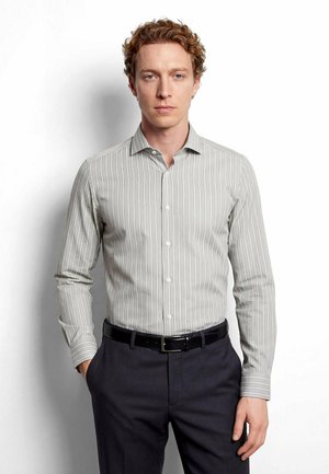 Mann mit lockigem Haar, der ein grau-weiß vertikal gestreiftes Hemd trägt, das in eine dunkle Hose mit einem schwarzen Ledergürtel gesteckt ist.