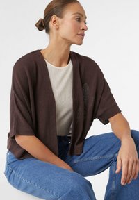 Bruine gebreide cardigan met korte mouwen, open ontwerp, gecombineerd met een lichtbeige textuurtop en blauwe denim jeans.