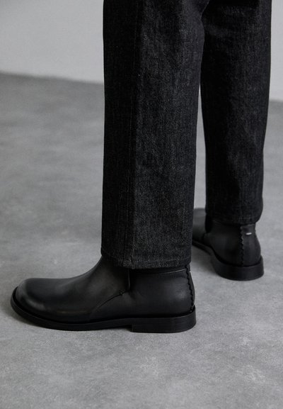Botas de tobillo de cuero negro con un acabado suave, puntera redonda y costuras en contraste. Combinadas con pantalones de mezclilla oscuros.