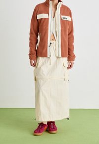 Brązowo-czerwony polar z kremowymi akcentami i zamkiem, połączony z długą beżową spódnicą cargo oraz bordowymi sneakersami z pomarańczowymi detalami.