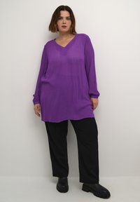 Haut à manches longues et décolleté en V en tissu violet audacieux, associé à un pantalon noir ample et des bottes noires épaisses. Texture lisse, coupe décontractée.